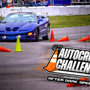 A blue sports car navigates an autocross course marked by orange cones, with a large "Autocross Challenge" logo overlayed in the foreground.