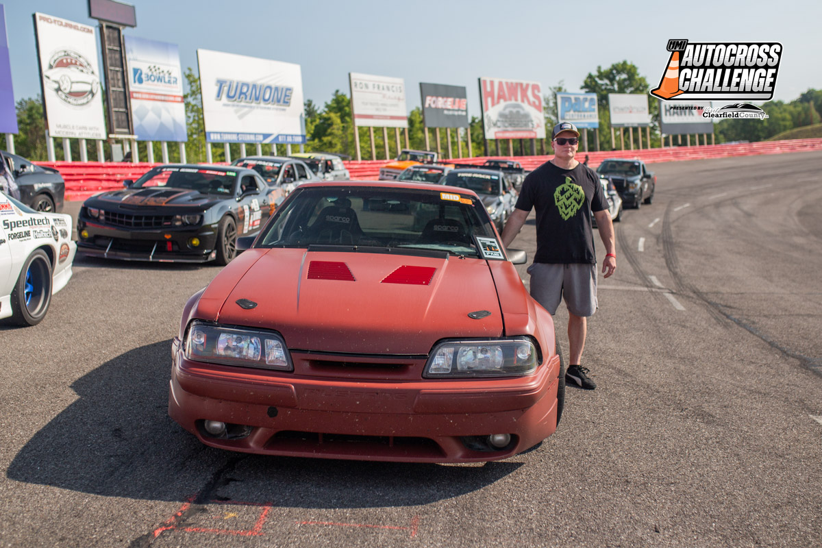 A person stands beside a red sports car on a racetrack, surrounded by other cars. The scene is part of the UM Autocross Challenge, as indicated by the banner in the background.