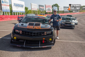 A man stands next to a black sports car at an autocross event, with several other cars lined up on a racetrack in the background. The event banner says "Autocross Challenge" on the right side.