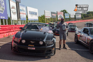 A man stands next to a black Ford Mustang holding a plaque at the Grassroots Motorsports Autocross Challenge event. The car and the track are adorned with various sponsor logos and decals.