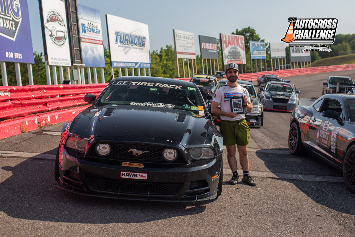 A person stands next to a black race car holding a trophy, with several other cars and a red barrier in the background. The scene is part of the Optima Autocross Challenge event.