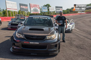 A man stands beside a black race car on a racetrack, holding a plaque. The car features various decals, and two other cars are seen in the background. An "Autocross Challenge" banner is visible.