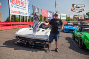 A man stands smiling next to a white sports car holding a flag and a trophy at an autocross event, with several other race cars parked in the background.