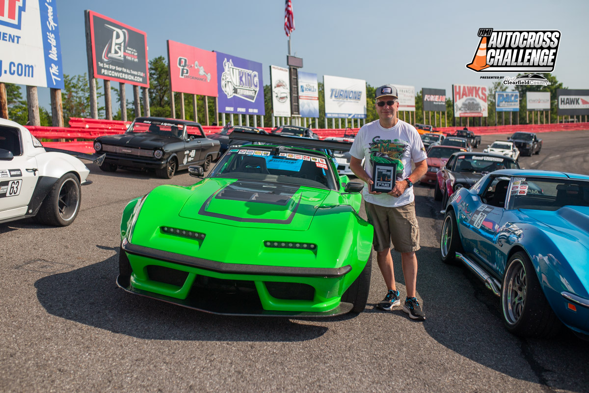 A man stands next to a bright green car holding a trophy, surrounded by other cars on a racetrack, with signs and an American flag in the background.