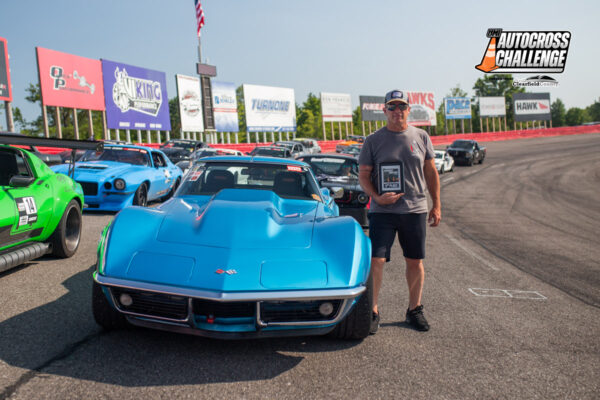 A person stands next to a blue vintage car on a racetrack, holding a plaque. Other cars and sponsor banners are visible in the background. The setting appears to be an automotive event.