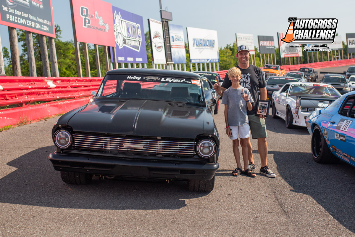 Two individuals stand next to a black vintage car on a racetrack during the Autocross Challenge event. Racing cars and sponsor banners are visible in the background.