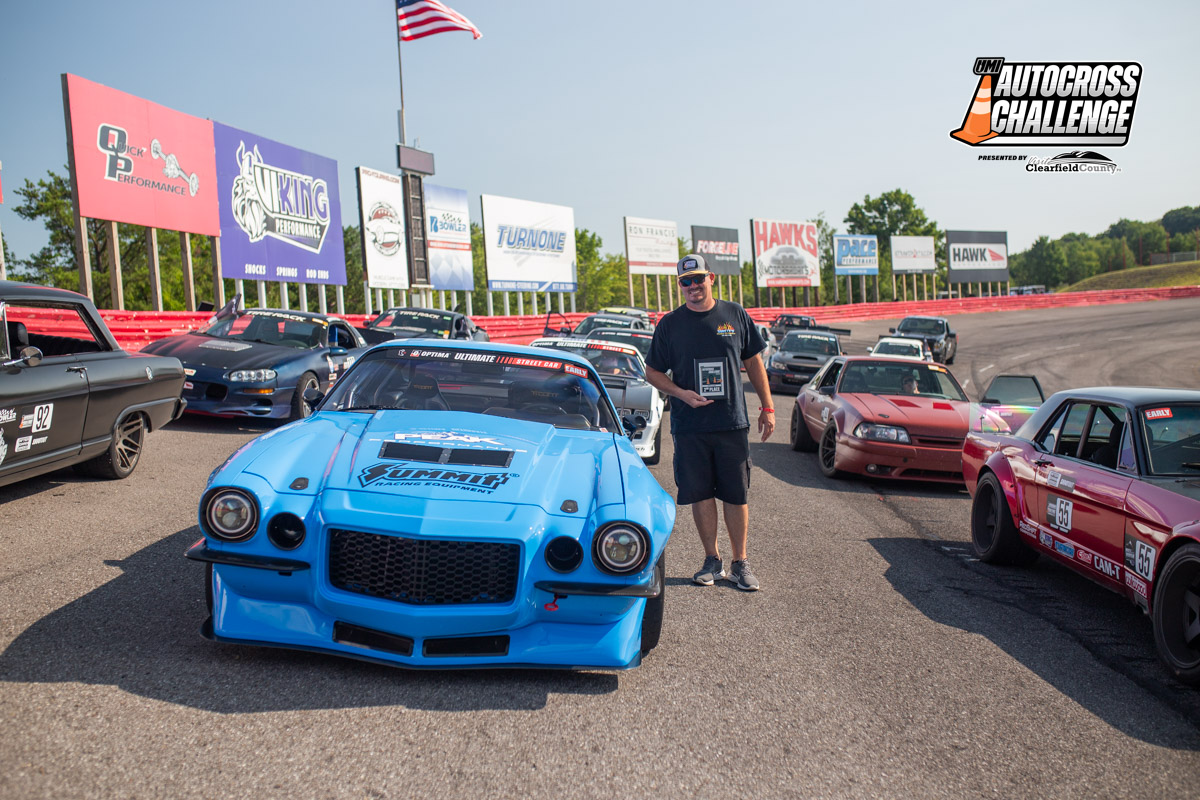 A man stands holding a plaque beside a blue race car, surrounded by other race cars, with various sponsor banners visible in the background, under a clear sky. The image is labeled: "Autocross Challenge.