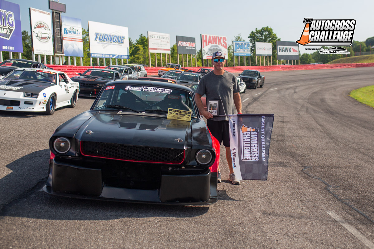 A man stands beside a black race car on a racetrack, holding a flag and a plaque. Several other race cars are parked in the background, and a logo for the Autocross Challenge is visible.