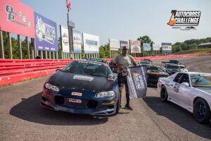 A man stands next to a black sports car on a racetrack holding a flag with the "Autocross Challenge" logo. Other cars and sponsor billboards are visible in the background.