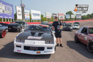 A person stands smiling next to a race car at an autocross challenge event. Other cars and various sponsor banners are visible in the background.