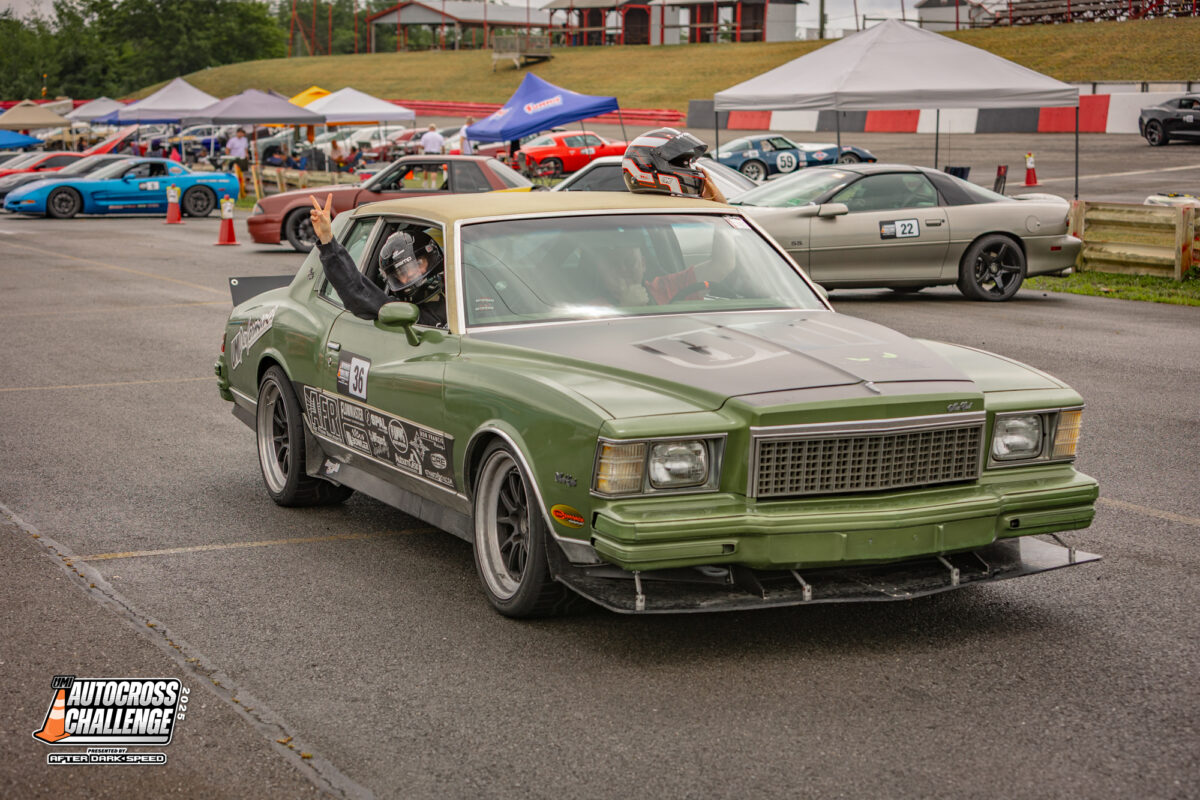 A green classic car with racing modifications is driven by a helmeted driver at an autocross event, with other cars and tents visible in the background.