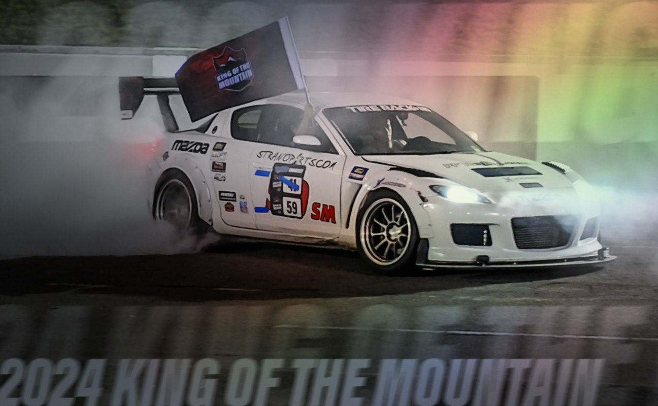 A white car with the number 59 and sponsor decals drifting on a racetrack, producing smoke. The text "2024 King of the Mountain" is partially visible at the bottom.