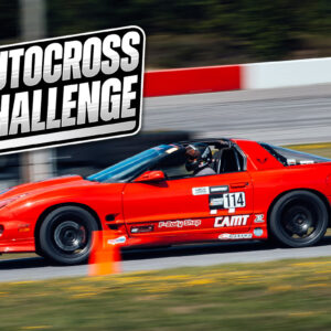 A bright red sports car with the number 114 races on a track at the UMI Autocross Challenge. An orange traffic cone is positioned on the track beside the car.