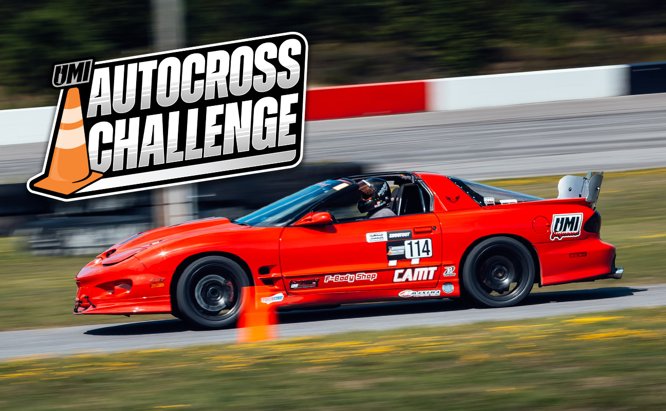 A bright red sports car with the number 114 races on a track at the UMI Autocross Challenge. An orange traffic cone is positioned on the track beside the car.