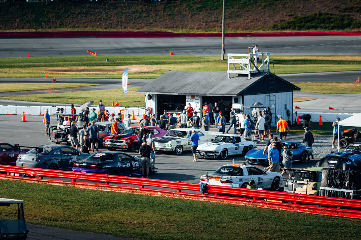 A group of people gather around various sports cars parked in front of a small building at a racetrack, with other vehicles and a grassy area in the background.