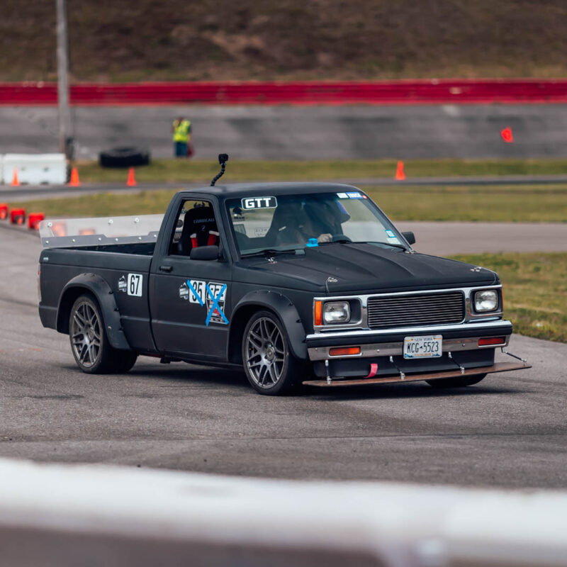 A black modified pickup truck, numbered 67, is seen racing on a track with red barriers and safety cones in the background. A person in a safety vest is visible in the distance.