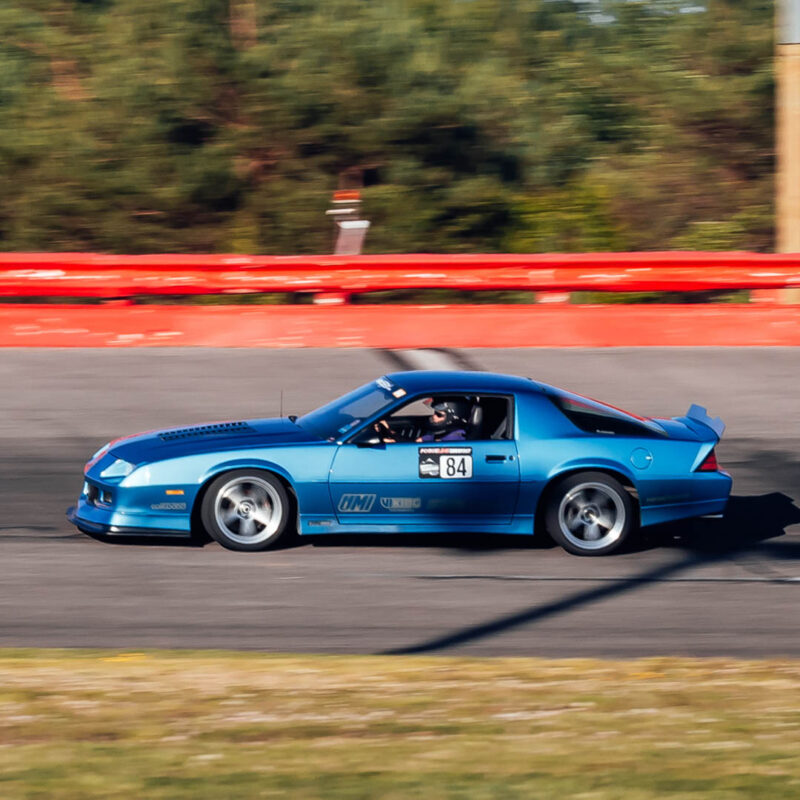 A blue sports car with the number 84 on the door is driving quickly on a racetrack. The driver is visible wearing a helmet. The background features a red barrier and blurred greenery.