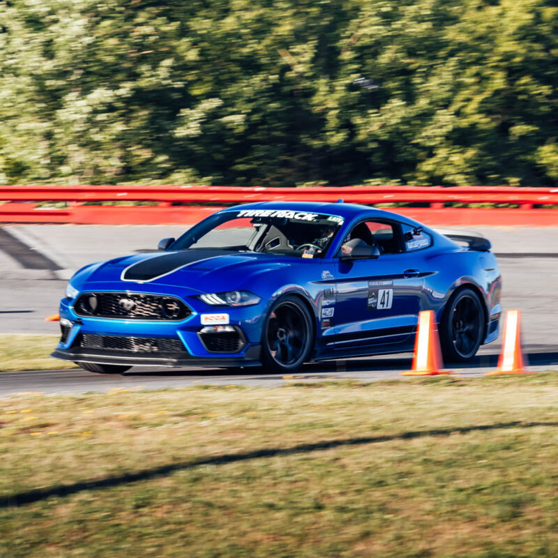 A blue sports car with the number 47 on its door races on a track, passing orange cones, with trees in the background.