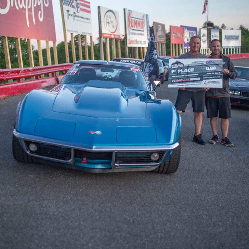 Two men are standing beside a blue sports car holding an oversized check for $1,500, marked as First Place. The background features racing-related signs and a racetrack.