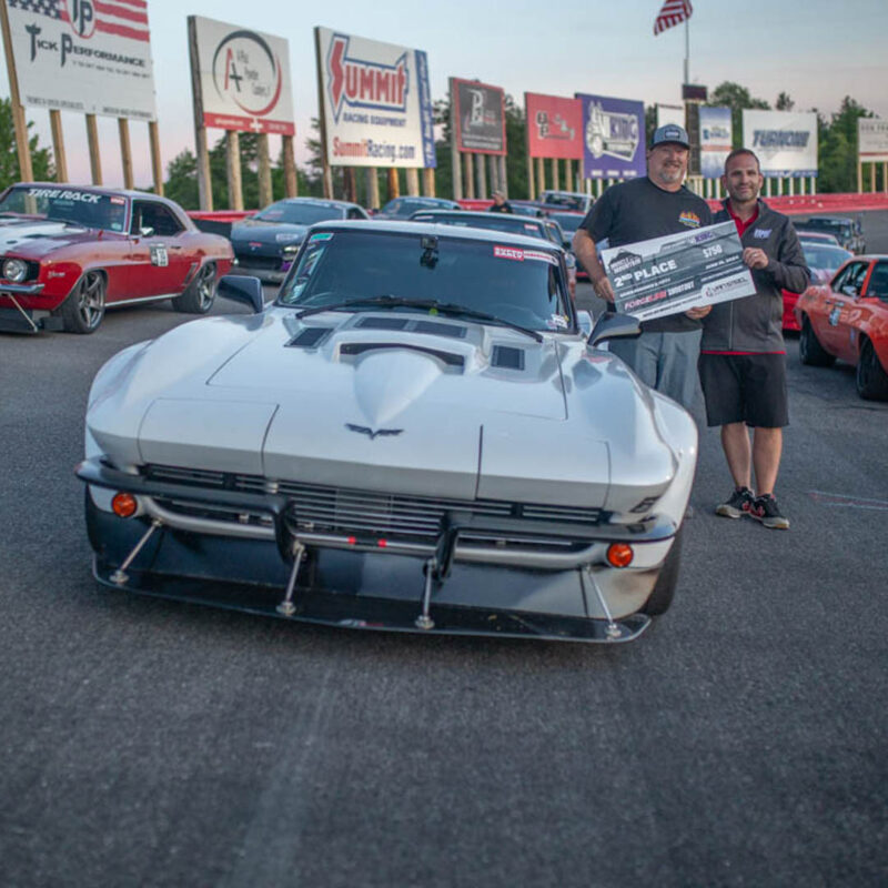 Two men stand in front of a silver sports car at a race track, holding a large check for winning second place. Other cars and sponsor banners are visible in the background.