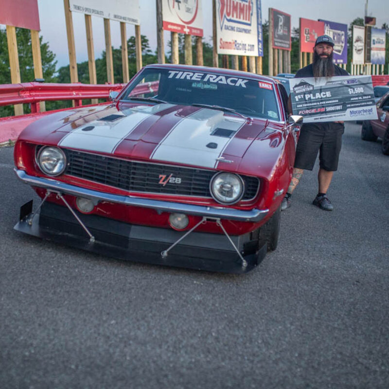 A man stands next to a red race car with a white stripe, holding a large check for $5,000 for winning 1st place at an automotive event.