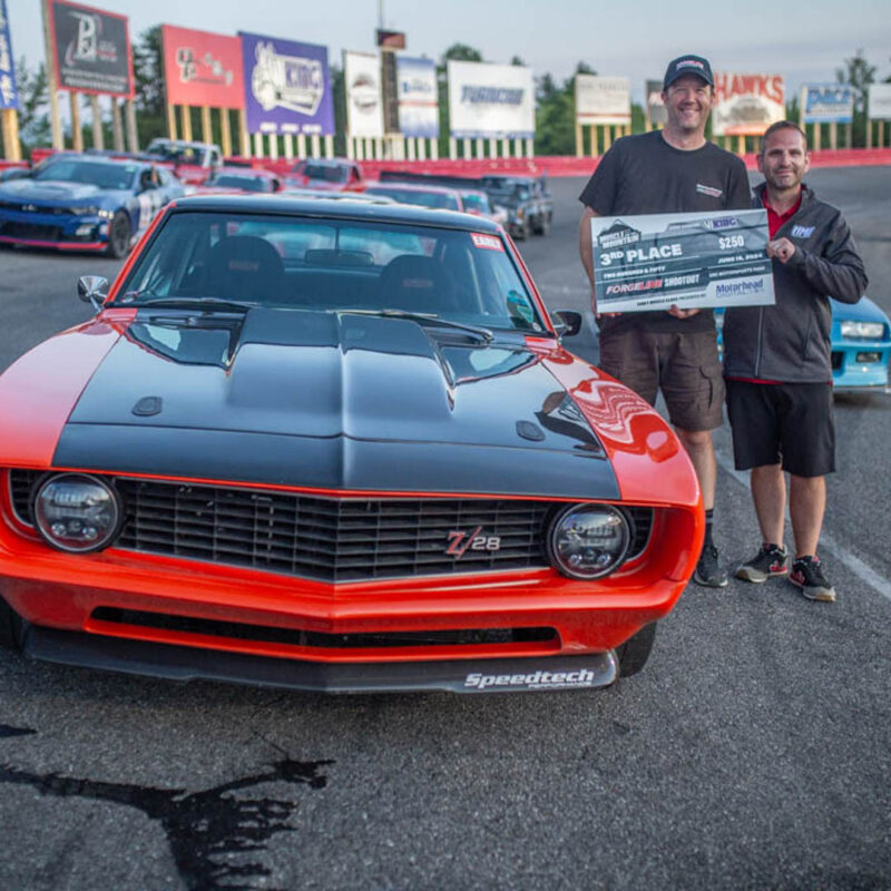 Two men stand beside a red Chevrolet Camaro Z28 holding a large third place check. The car is parked on a racetrack with several other cars in the background.