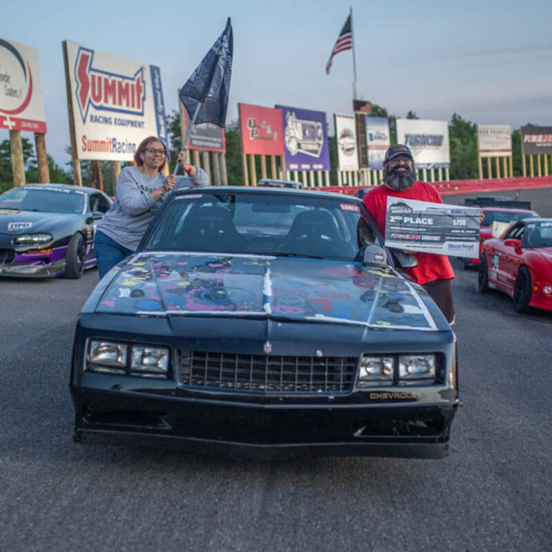 Two people stand beside a black Chevrolet car on a racetrack, holding flags and a 2nd place prize poster. Other cars and advertisement boards are visible in the background.