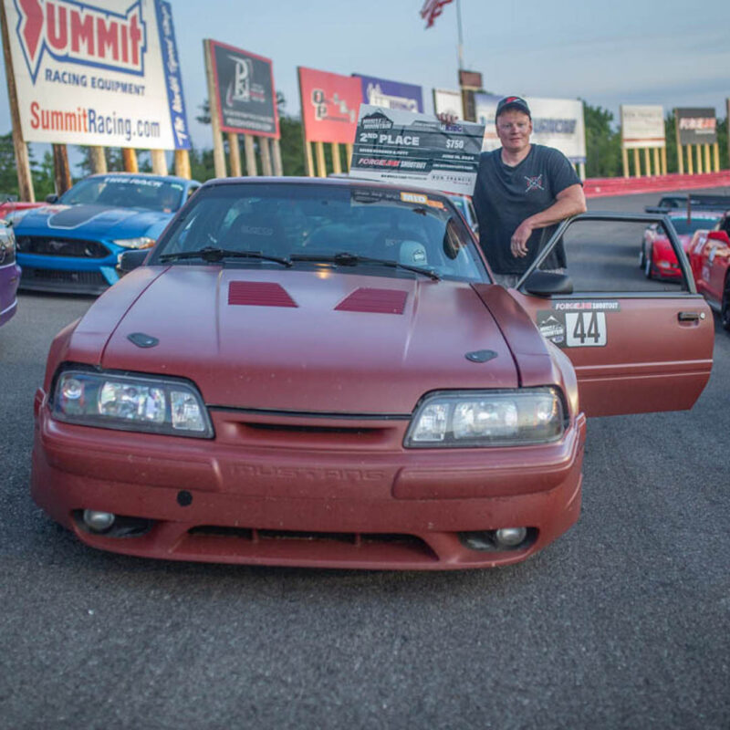 A person stands next to an open car door, leaning on a red sports car with racing decals, including the number 44, in a racetrack setting with several parked cars and advertising banners in the background.