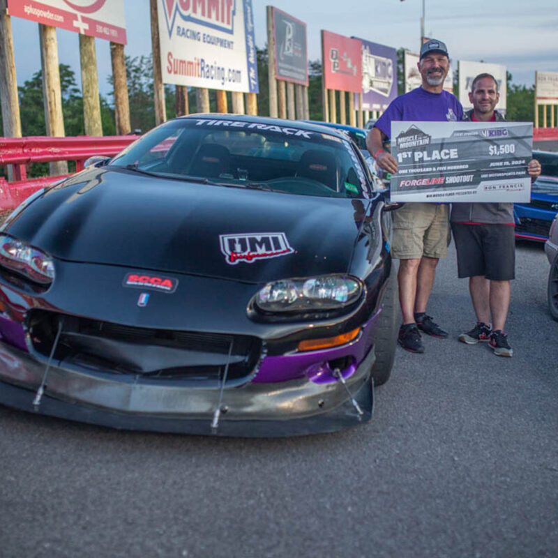 Two people standing in front of a black race car hold a large first-place check for $1,500. The car features various decals and a prominent front bumper modification.