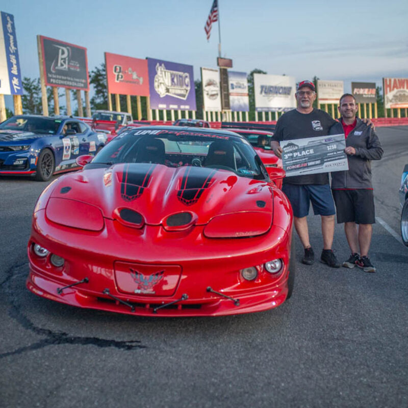 Two men stand next to a red sports car holding a large check for third place. They are at a racetrack with advertising banners in the background.