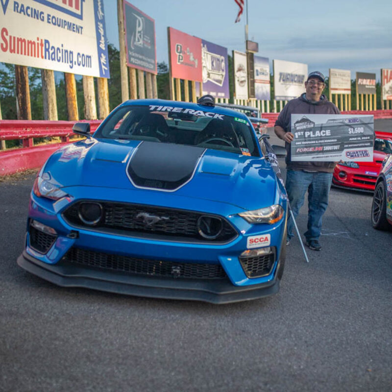 A person stands next to a blue sports car on a race track, holding a large 1st place prize check for $1,500.