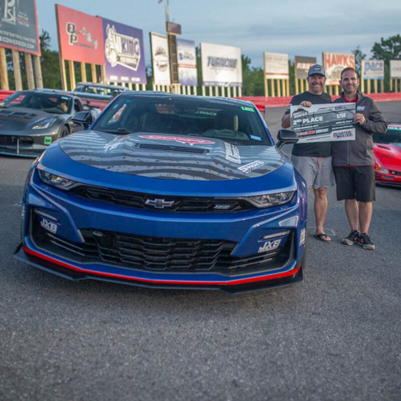 Two men stand next to a blue Chevrolet racing car holding a check for "2nd Place" on a racetrack surrounded by other cars and advertisement banners.