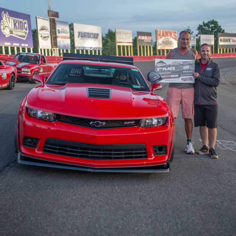 Two men stand beside a red Chevrolet Camaro on a racetrack, holding a 3rd place check.