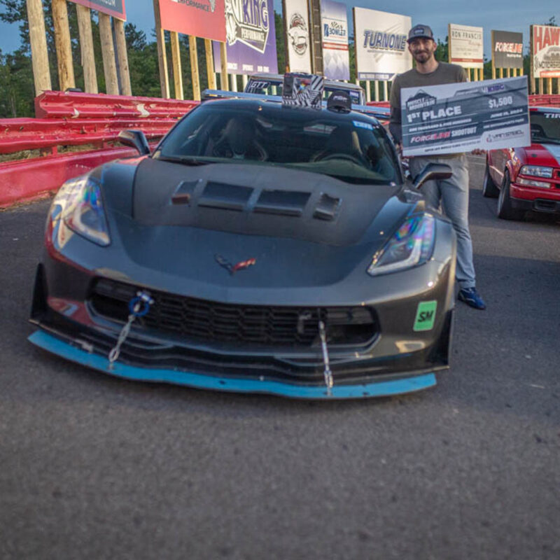 A person wearing a cap stands beside a sports car holding a large first-place check. Several cars and advertising banners are visible in the background.