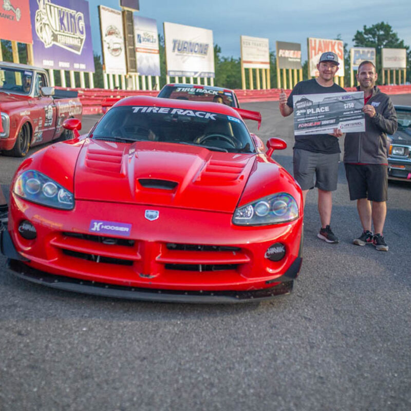 Two men standing on a race track hold a sign for a 2nd place achievement next to a red racing car, with several other vehicles and advertising boards in the background.