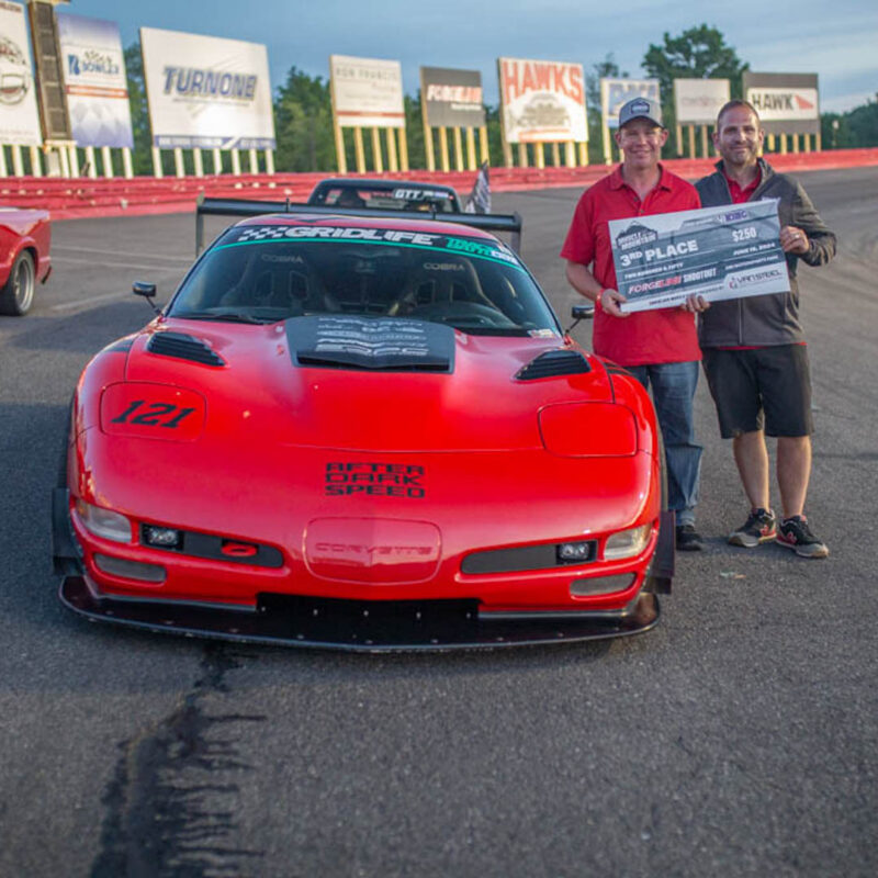 Two men pose next to a red sports car on a race track, with one holding a large check for a third-place finish. Racing barriers and sponsor banners line the background.