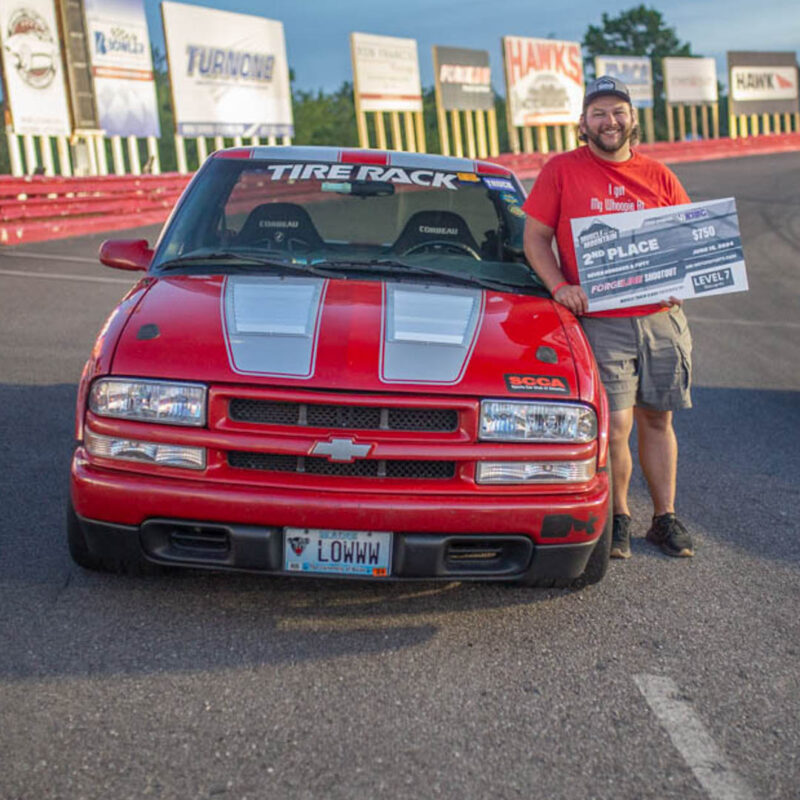 A person stands next to a red Chevrolet truck holding a prize check. The truck has racing decals, and the license plate reads "LOHWW." The background shows a race track with various advertising banners.