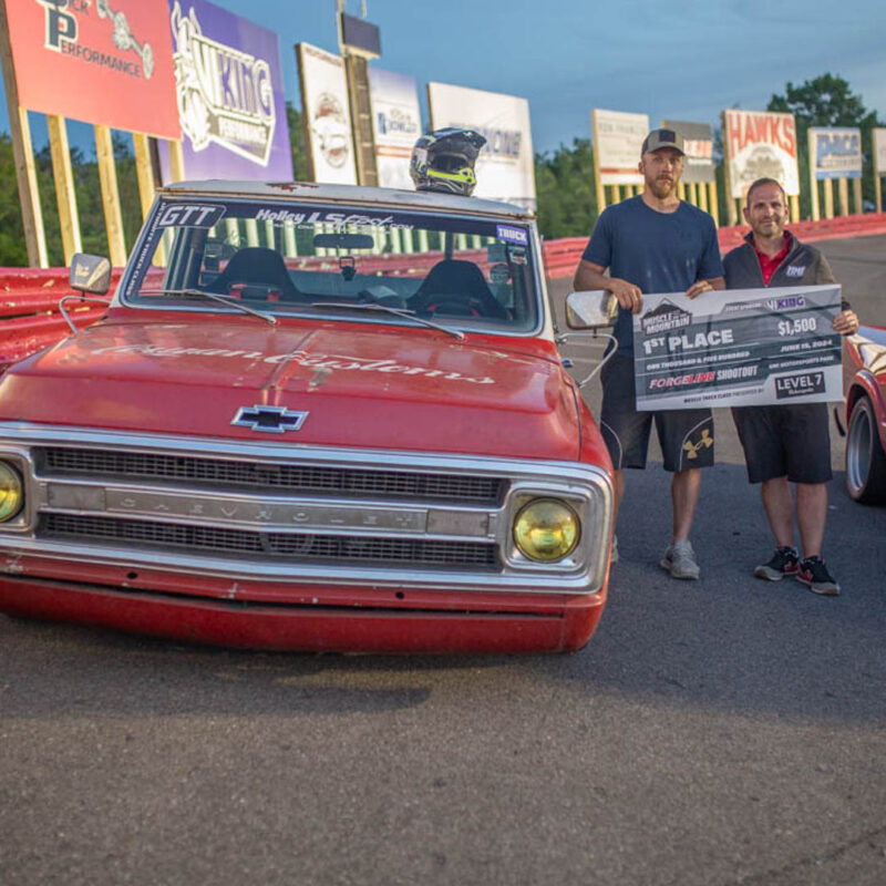 Two men holding a large check stand beside a classic red Chevrolet truck on a racetrack, surrounded by racing banners.