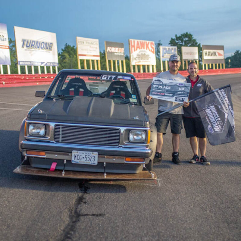 Two people stand next to a car in front of a race track, holding an award placard and a flag. The car has a license plate with the number "KCG-5523" and multiple sponsor logos are visible in the background.