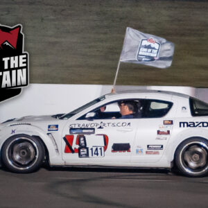 A driver waves a flag out of a mazda race car with "king of the mountain" branding in the background.