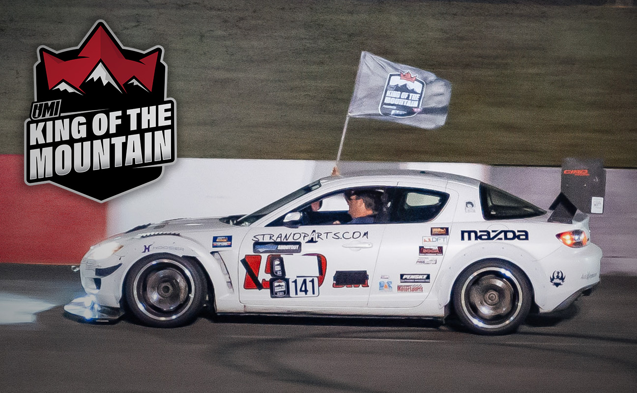 A driver waves a flag out of a mazda race car with "king of the mountain" branding in the background.