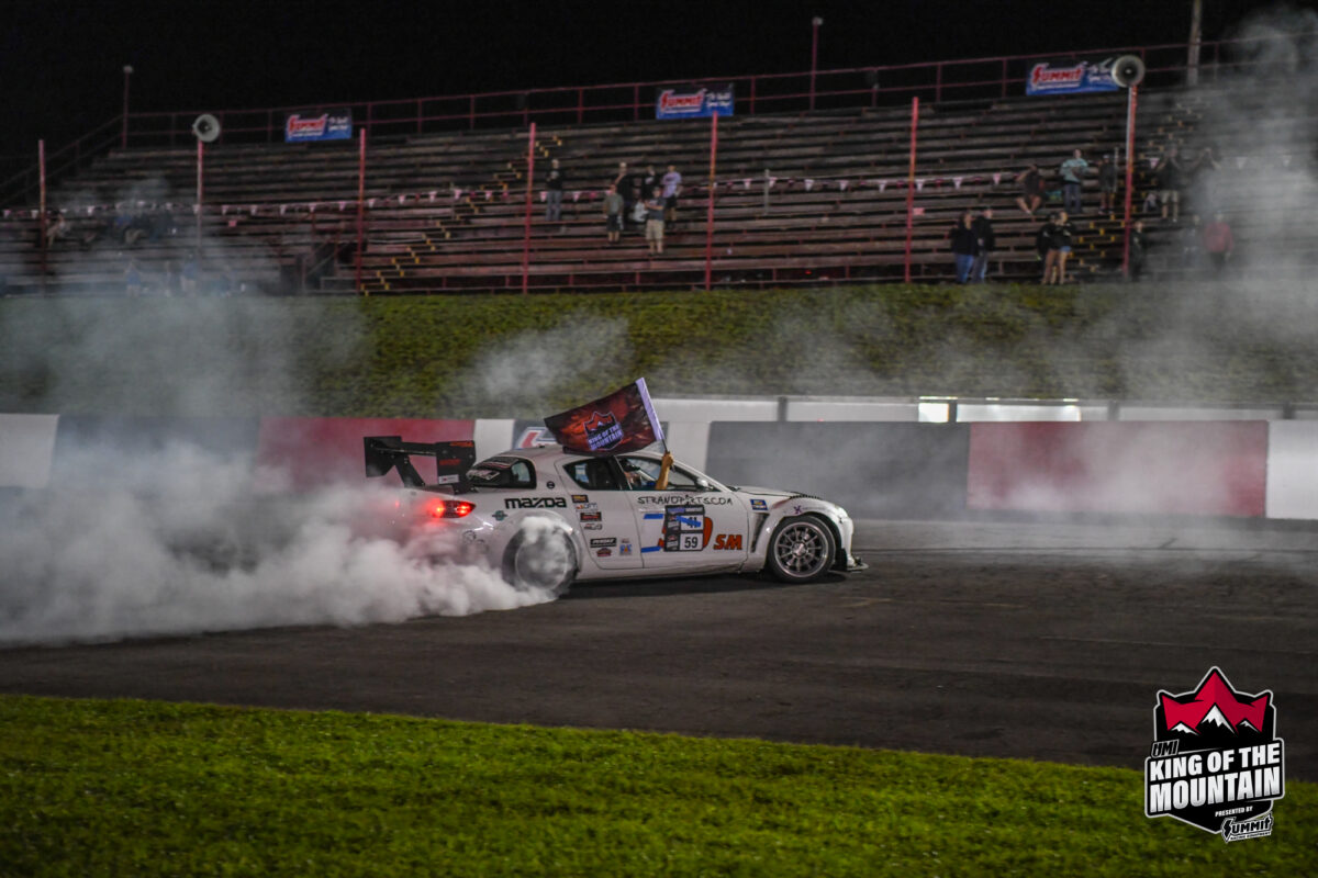 A Mazda race car drifts on a track, producing smoke from its tires. The "King of the Mountain" event logo appears in the bottom right corner. Stands in the background have few spectators.