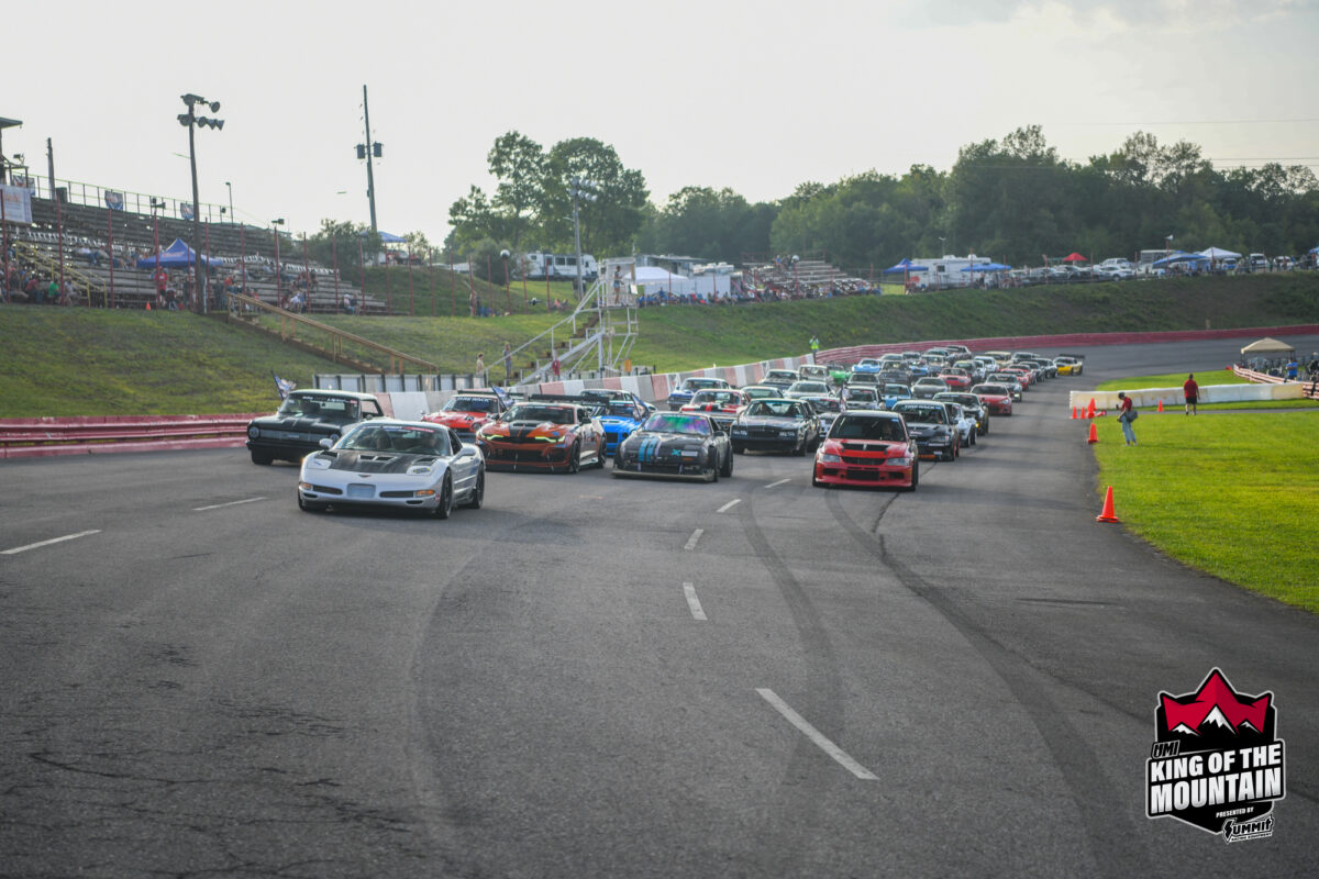 A large group of race cars, including various models and colors, are tightly packed and racing on a paved track at King of the Mountain. Spectators can be seen in the stands in the background.