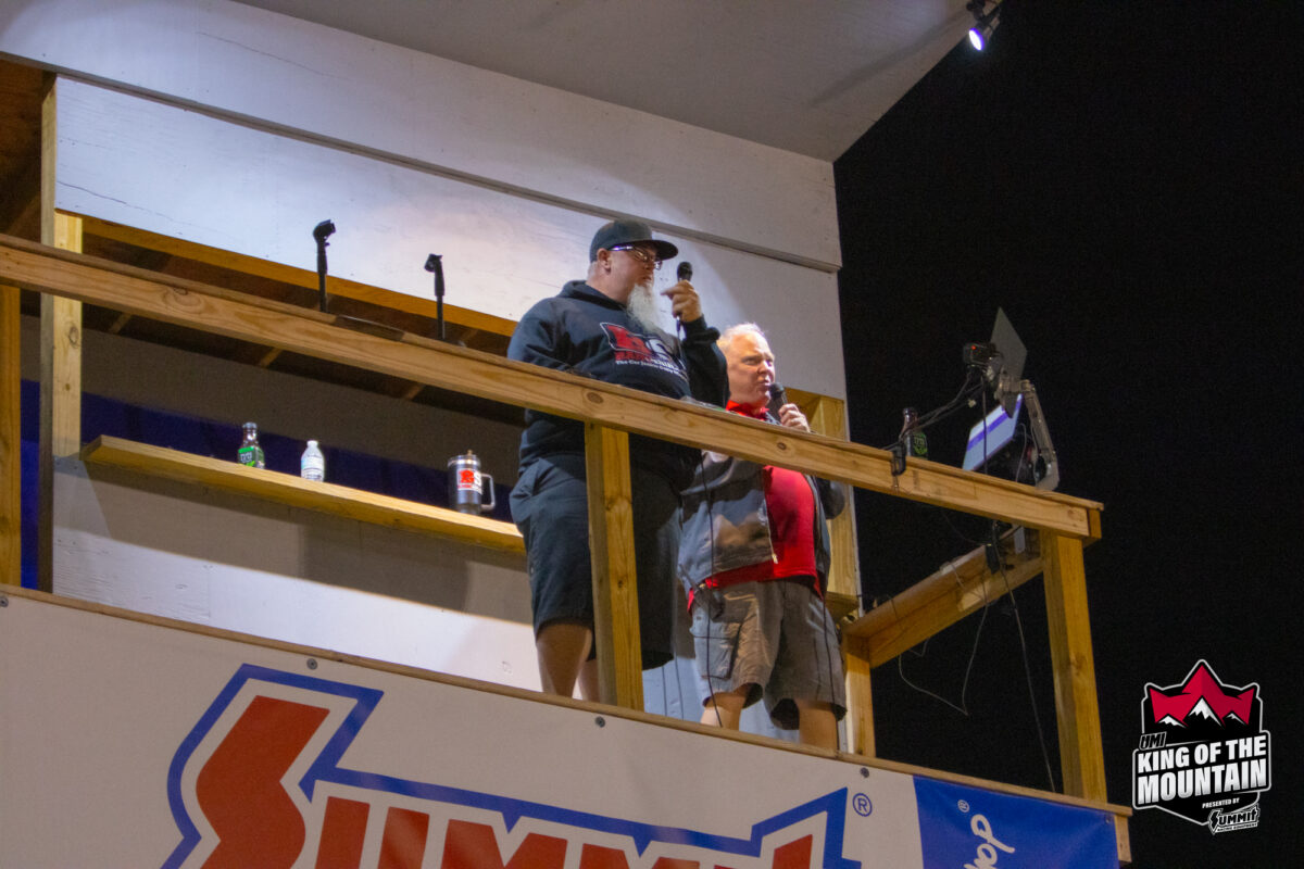 Two individuals are standing on an elevated platform holding microphones at what appears to be an event, with a "Summit" sign and a "King of the Mountain" logo visible below them.