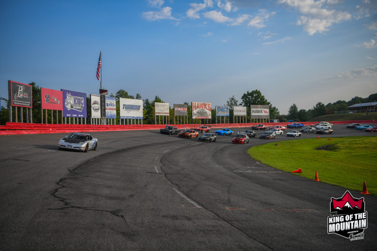 A group of cars racing on a paved track with numerous billboards and an American flag in the background. The "King of the Mountain" logo is displayed in the bottom-right corner.