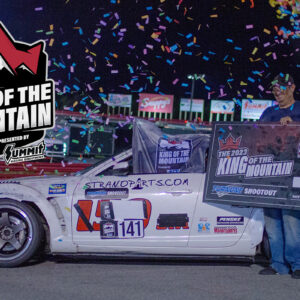 A man stands beside a white race car, holding a large check for $15,000 at the umi king of the mountain event, with confetti in the air.