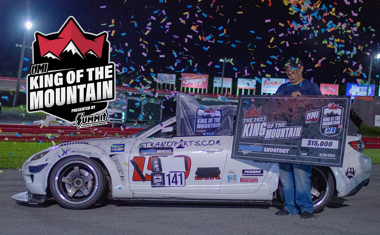 A man stands beside a white race car, holding a large check for $15,000 at the umi king of the mountain event, with confetti in the air.