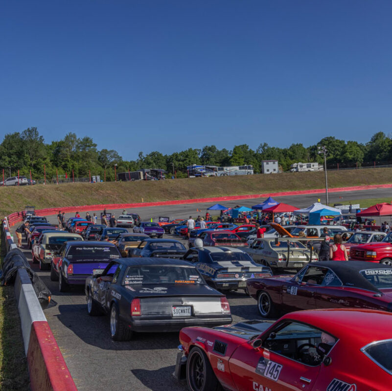 A lineup of various parked sports cars on a racetrack, with drivers and teams preparing under tents. The scene is set against a backdrop of trees and a clear blue sky.