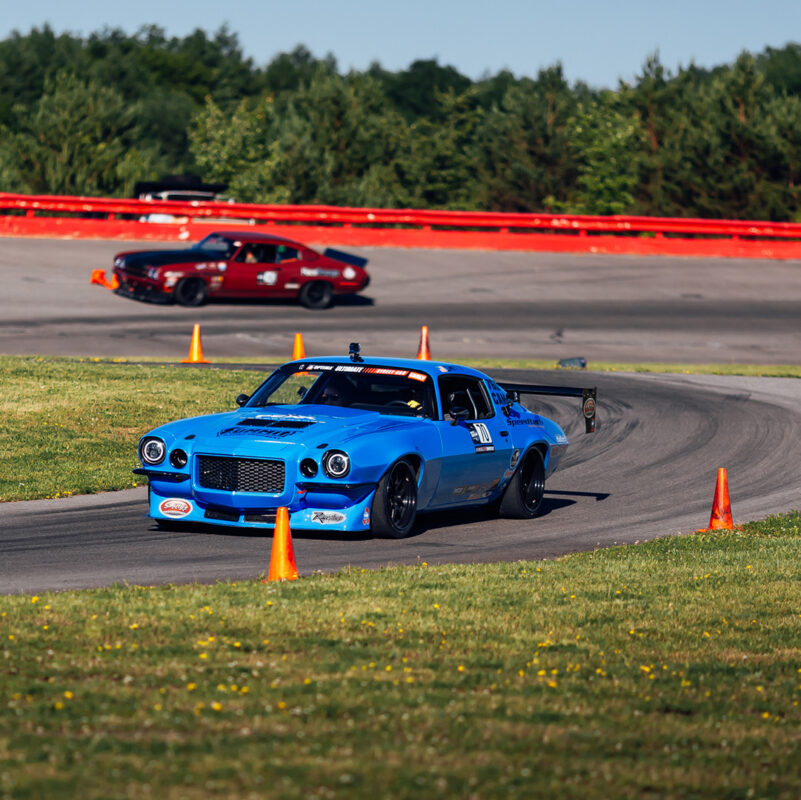 A blue racing car takes a corner on a race track, followed at a distance by a red racing car. Orange traffic cones line the track, and green trees are visible in the background.