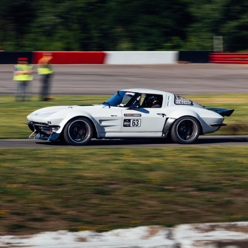 A white race car with the number 63 on the side drives on a racetrack with blurred motion, indicating high speed. Radio control operators and safety barriers are visible in the background.